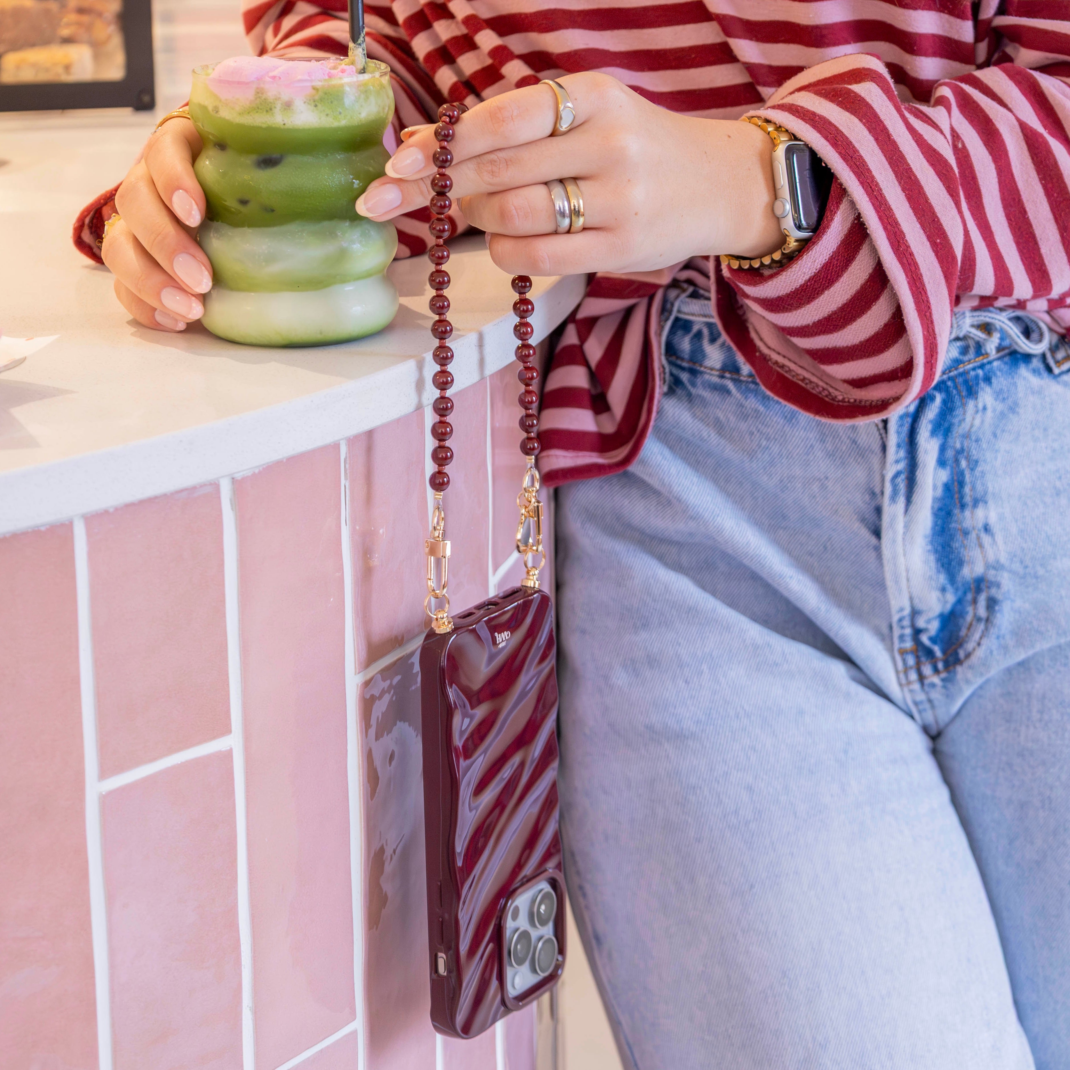 Vrouw met bordeauxrode kralen telefoonkoord en bijpassend hoesje, gouden haakjes, aan een roze tegelbar.