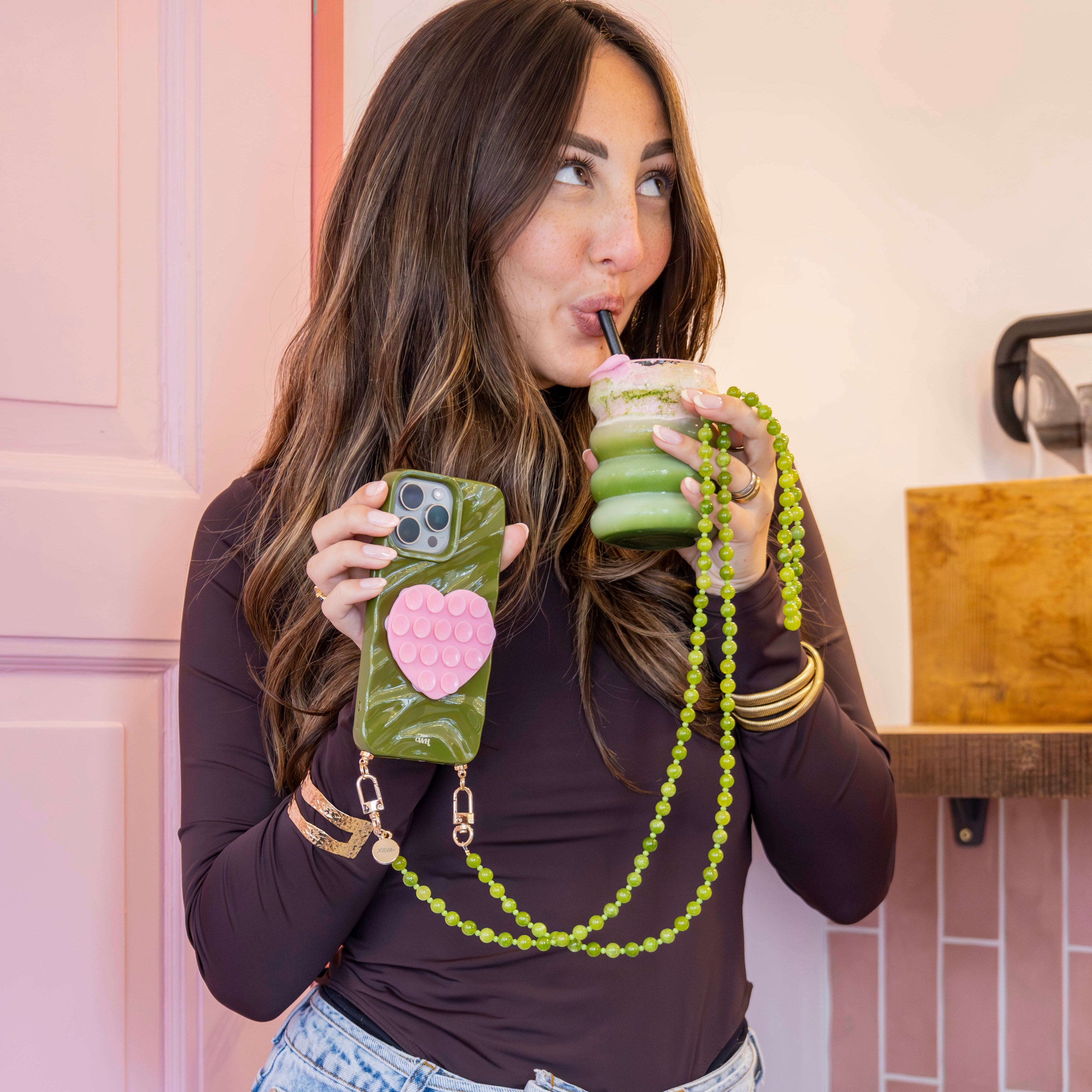Vrouw met lange Deep Olives telefoonkoord en bijpassend hoesje met roze hart, drinkt een groene drank.