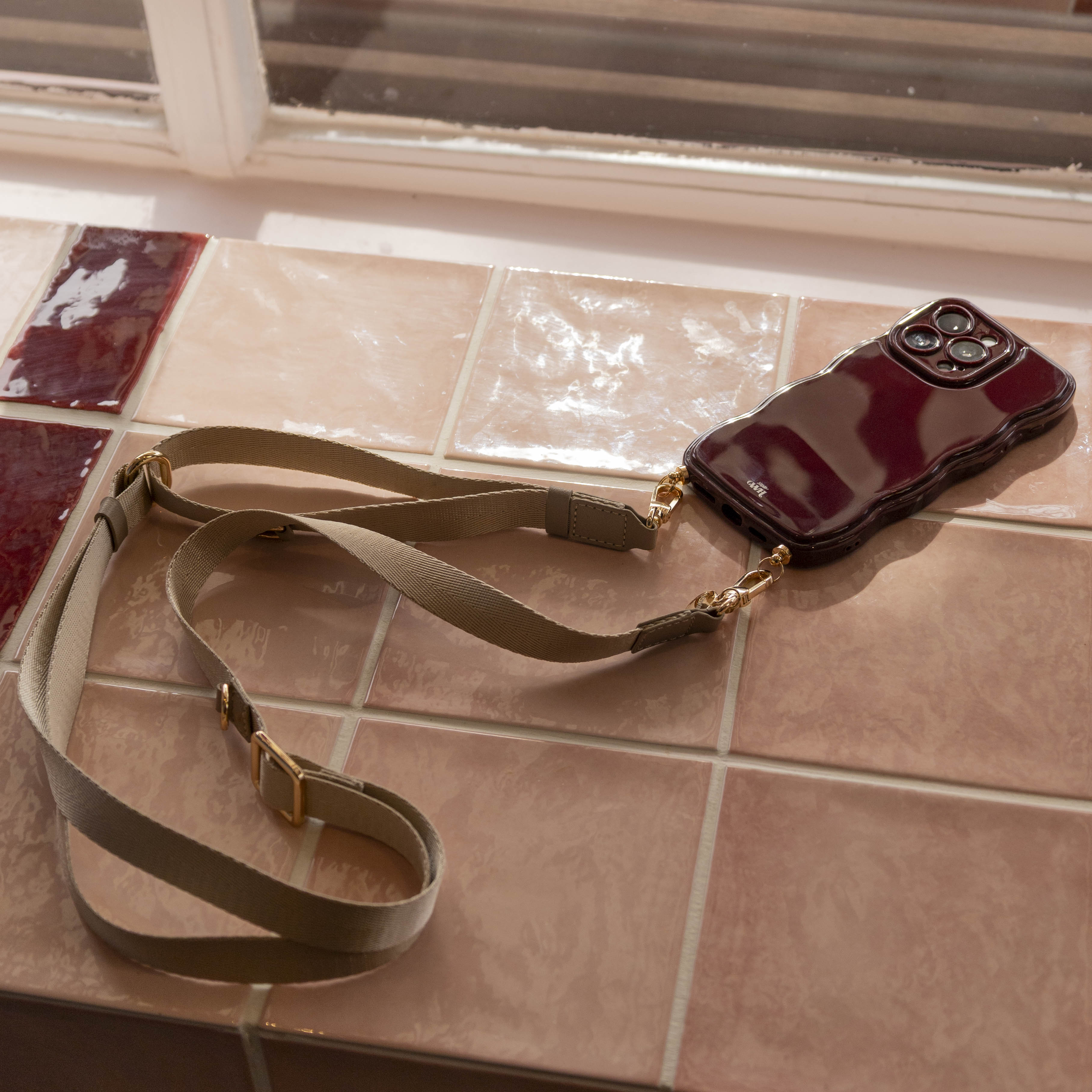 Beige lanyard strap met gouden clips bevestigd aan een glanzend bordeauxrood telefoonhoesje op roze tegels.