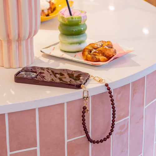 Kort Burgundy Muse koord met gouden haakjes en bordeauxrode kralen, bevestigd aan een golvend telefoonhoesje.