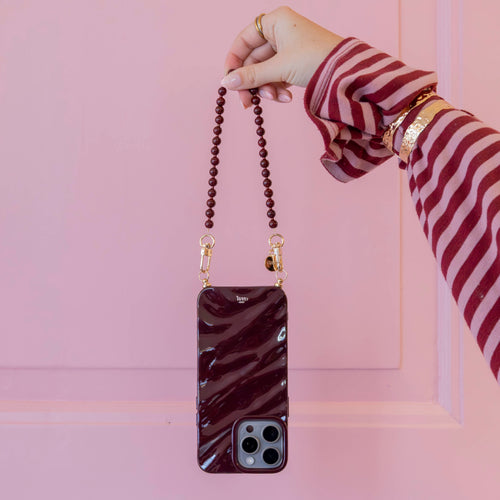 Vrouwenhand houdt een korte, glanzende bordeauxrode telefoonhoes met bijpassend kralenkoord en gouden sluitingen vast.