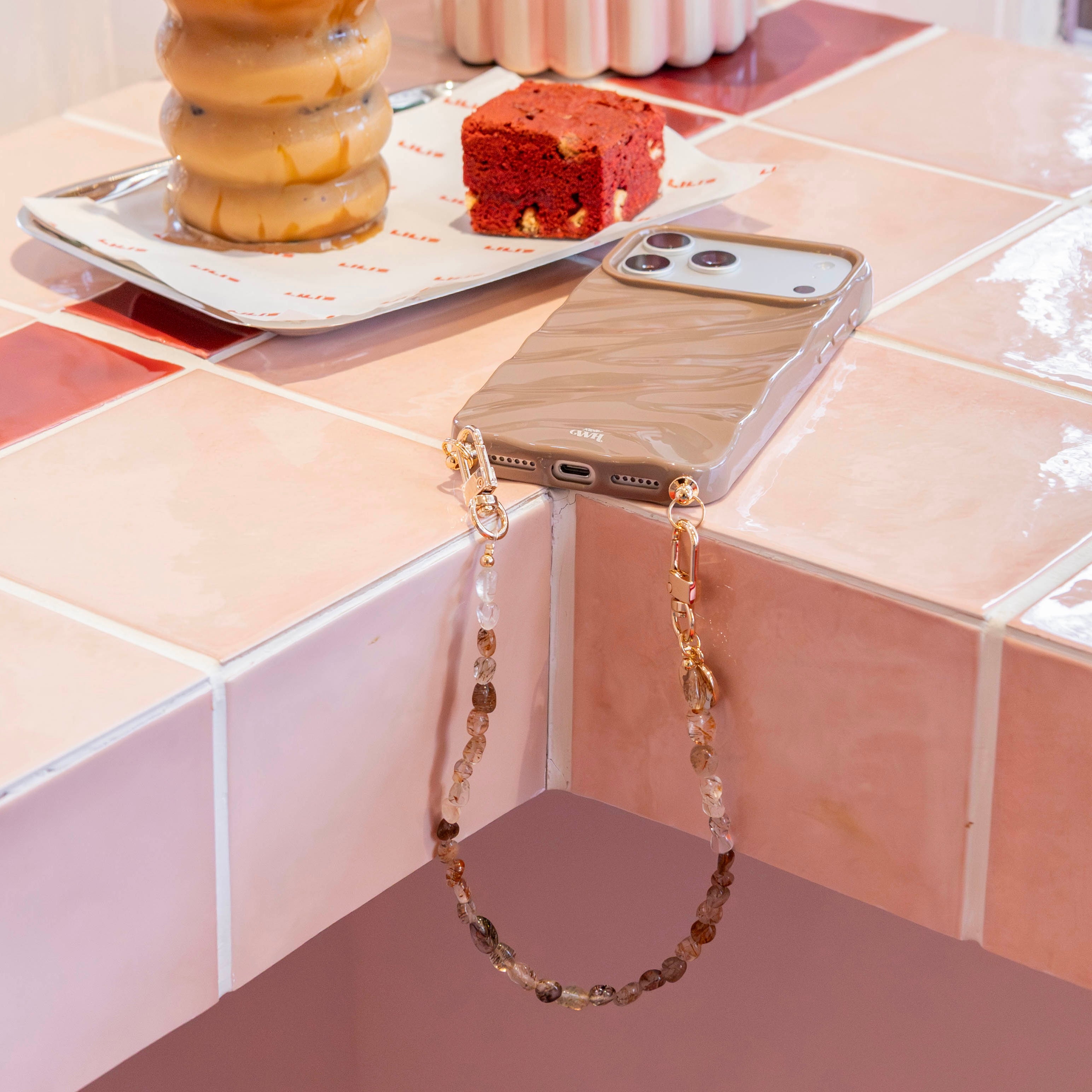 Kort Soft Taupe koord met gouden haakjes en kralen, bevestigd aan een telefoonhoesje.