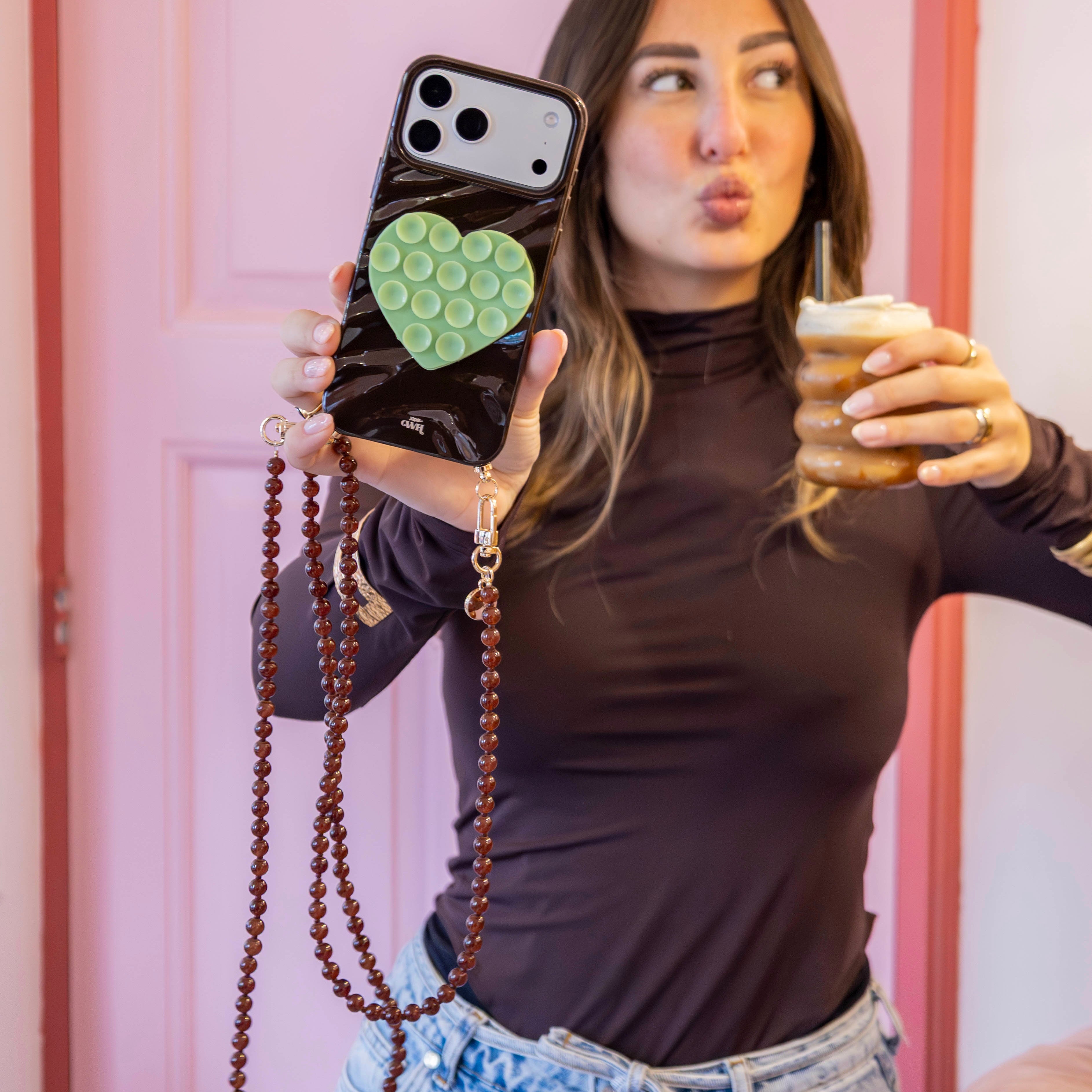 Vrouw toont glanzende bruine iPhone 13 Glaze Cord Case met groen hart en lang bruin Sugar koord.