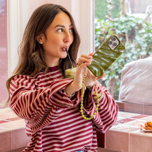 Vrouw drinkt uit een glas terwijl ze een iPhone 17 met glanzende Deep Olives Glaze Cord Case vasthoudt.