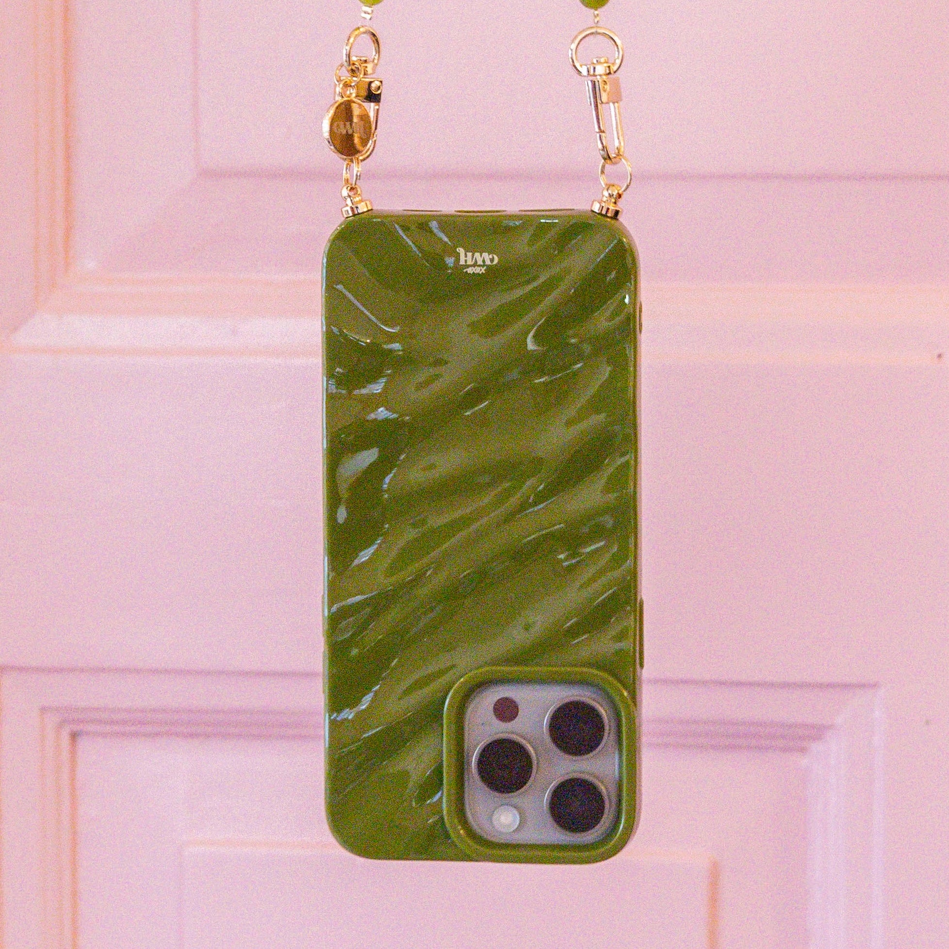 Glanzende groene iPhone 14 Glaze Cord Case met marmerlook en gouden hardware, hangend voor een roze deur.