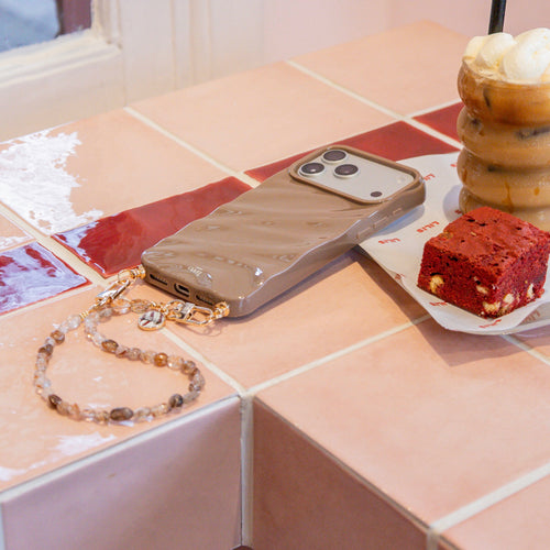 iPhone 17 Pro in Taupe Glaze Cord Case met Soft Taupe polskoord en gouden details op een tegelblad.