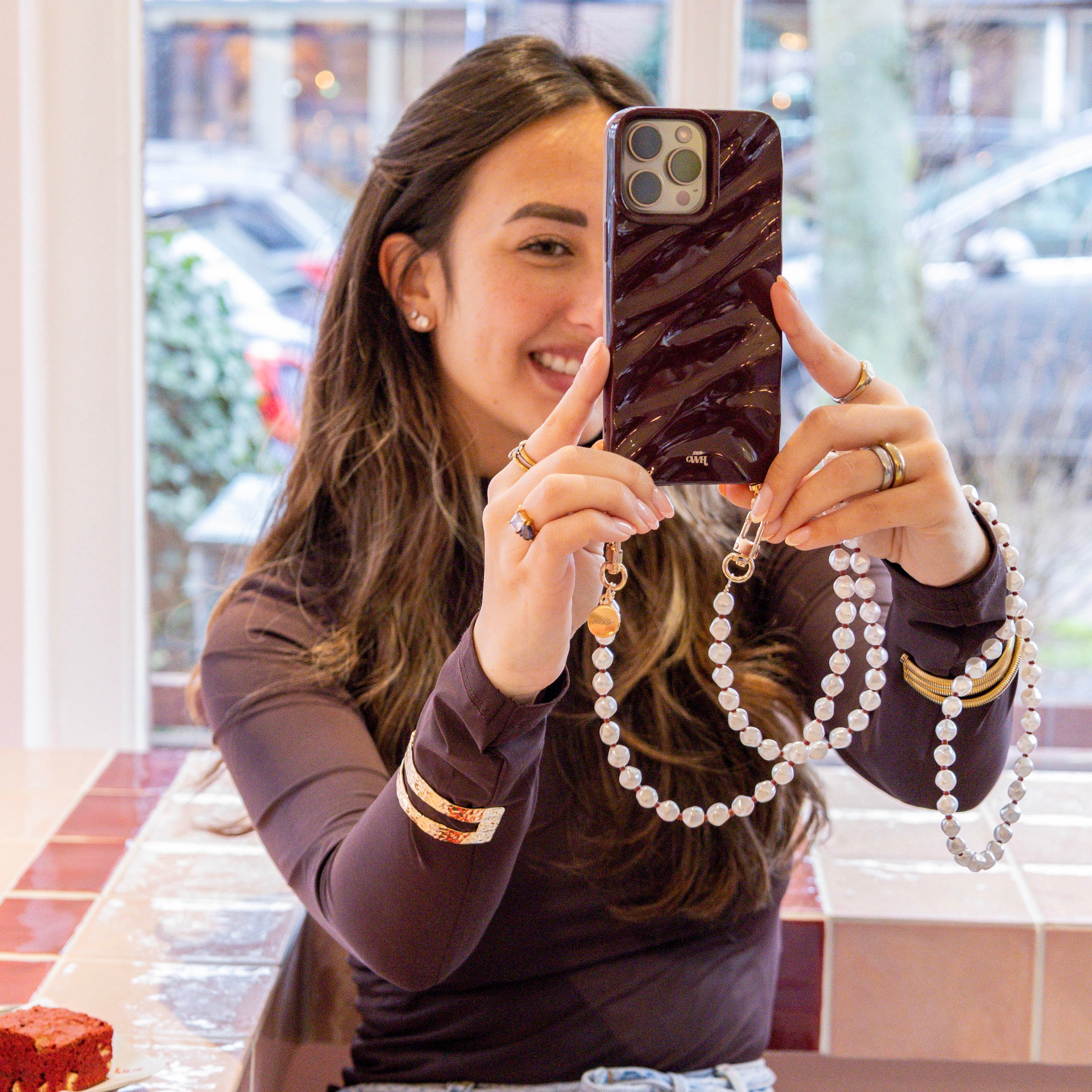 Vrouw toont glanzende bordeauxrode iPhone 15 Glaze Cord Case met lang Burgundy Pearl parelkoord.