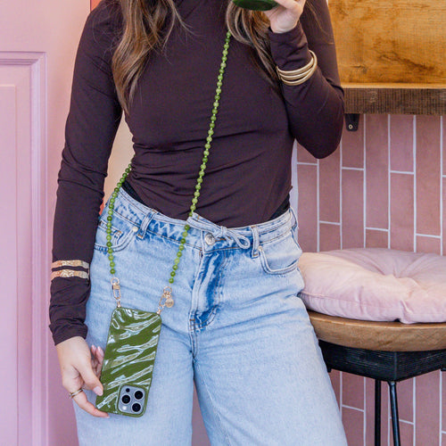 Vrouw draagt iPhone 17 Glaze Cord Case in Deep Olives met lang groen kralenkoord crossbody.