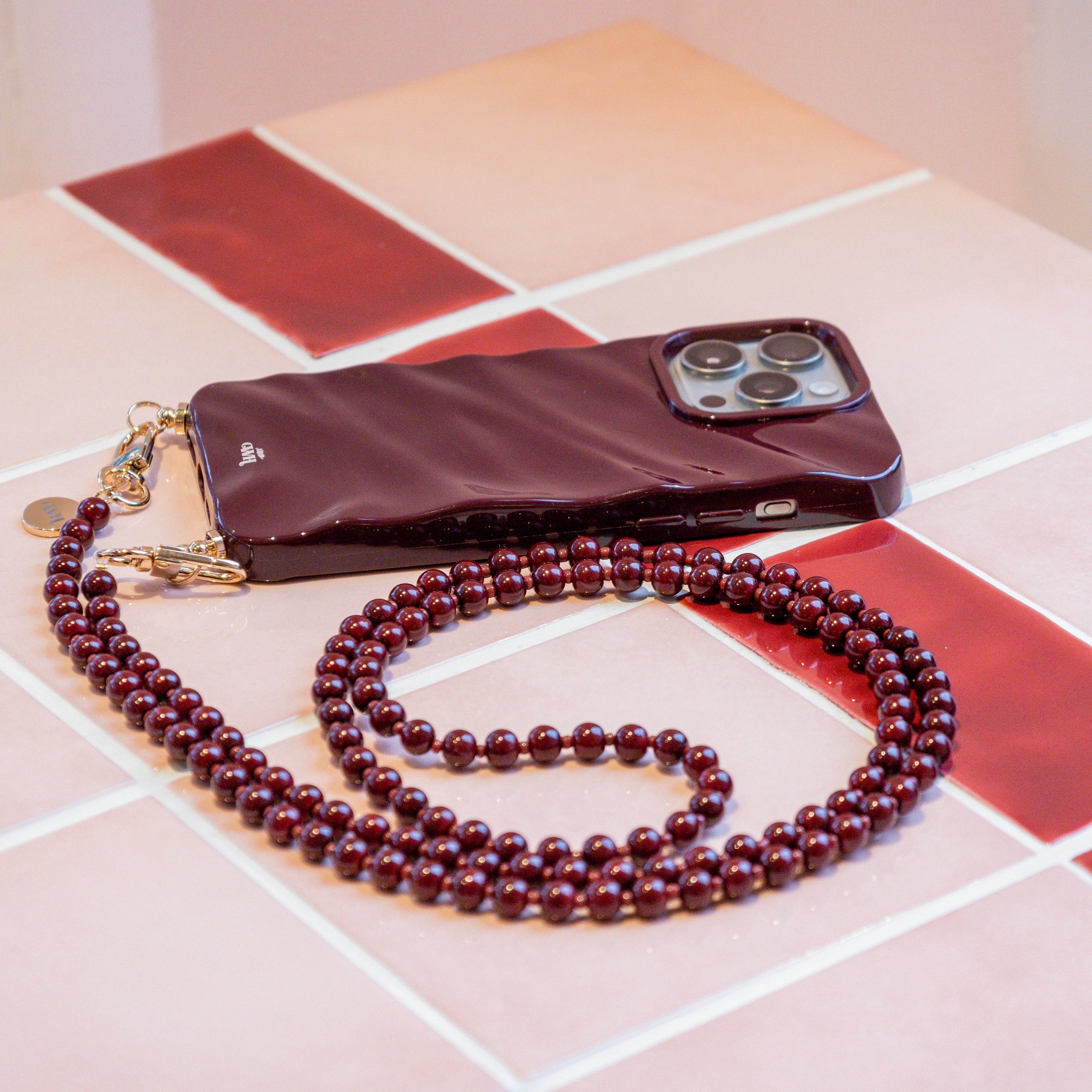 Glanzende bordeauxrode iPhone 15 Glaze Cord Case met bijpassend lang Burgundy Muse koord en gouden details.