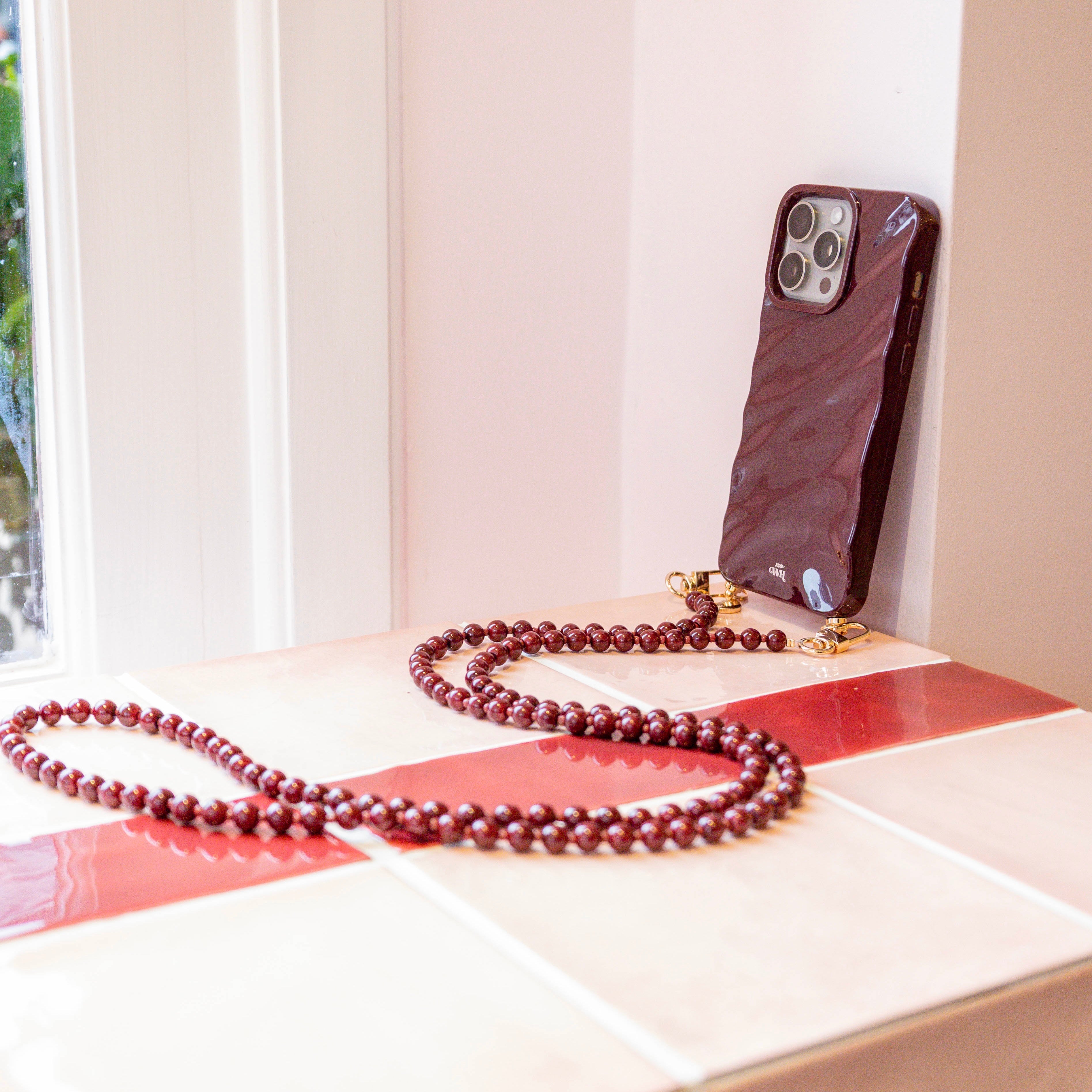 Glanzend bordeauxrode iPhone 15 Plus Glaze Cord Case met lang Burgundy Muse koord op een vensterbank.