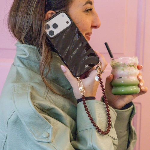 Vrouw houdt een iPhone 16 Pro Max met een glanzende bruine Glaze Cord Case en een bruine kralenketting vast.