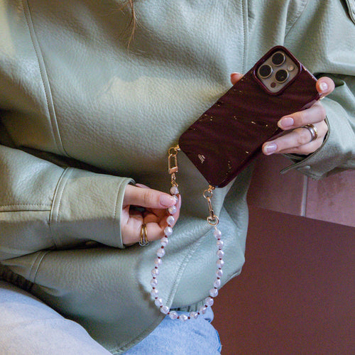 Hand houdt een glanzende bordeauxrode iPhone 16 Pro Max case met parelkoord en gouden accenten vast.