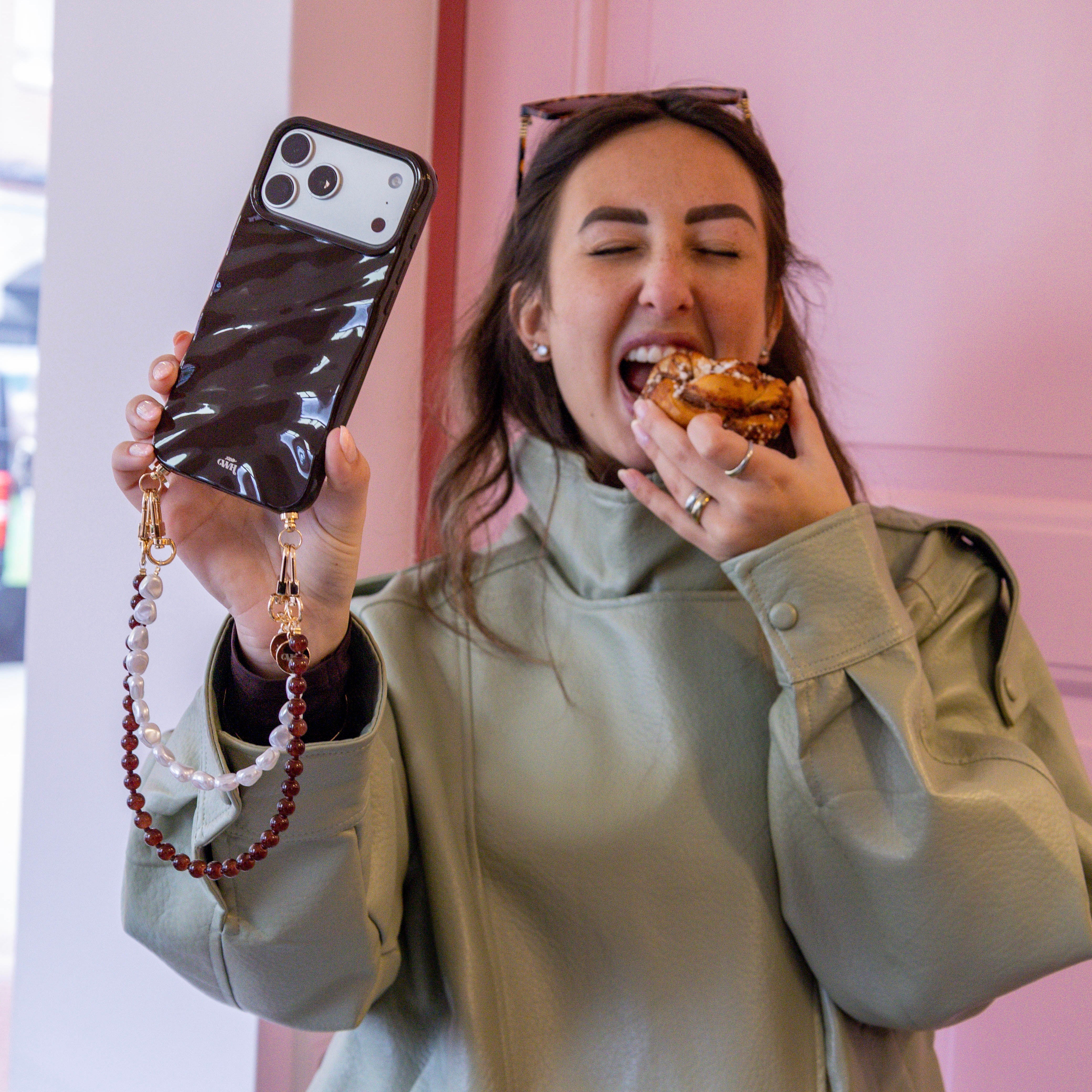 Vrouw met glanzend bruin iPhone 13 Pro Max hoesje met parel- en bruine kralenkoord, eet een kaneelbroodje.