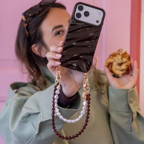 Vrouw toont glanzende bruine iPhone 13 Pro Max hoes met parel- en bruine kralenkoord.