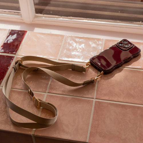Beige lanyard strap met gouden clips bevestigd aan een glanzend bordeauxrood telefoonhoesje op roze tegels.