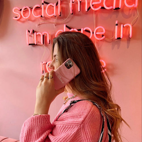 Vrouw met lang haar draagt roze trui en houdt een gepersonaliseerd roze telefoonhoesje met "selflove" tekst vast.