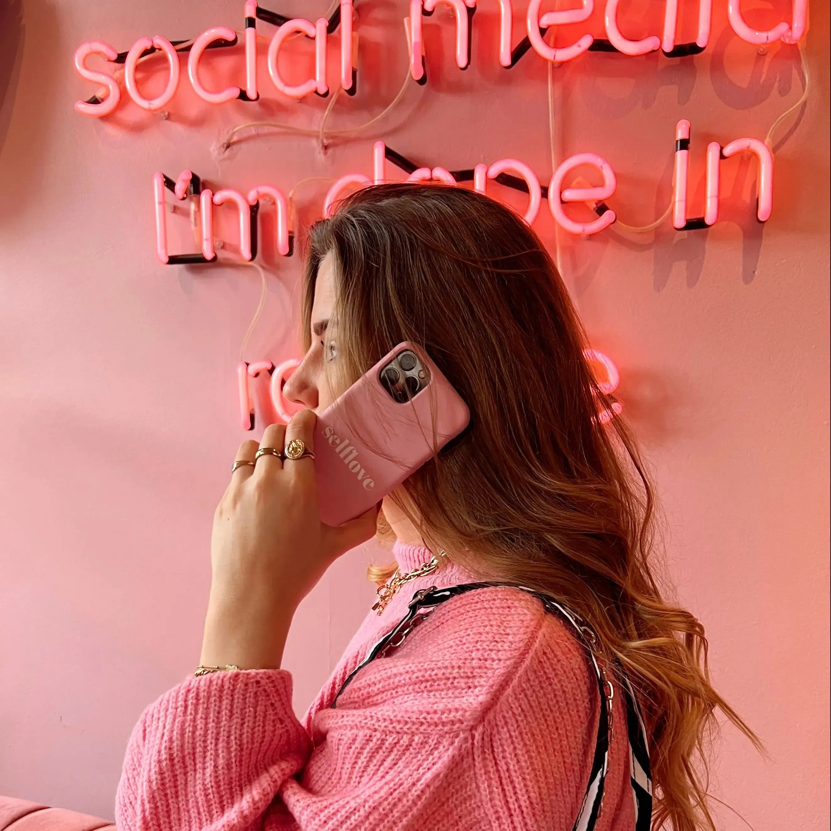 Vrouw met lang haar draagt roze trui en houdt een gepersonaliseerd roze telefoonhoesje met "selflove" tekst vast.