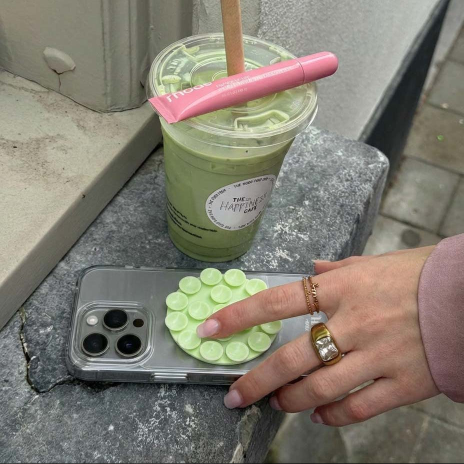 Ronde, lichtgroene siliconen sticker met zuignappen op een transparant telefoonhoesje, naast een groene drank.