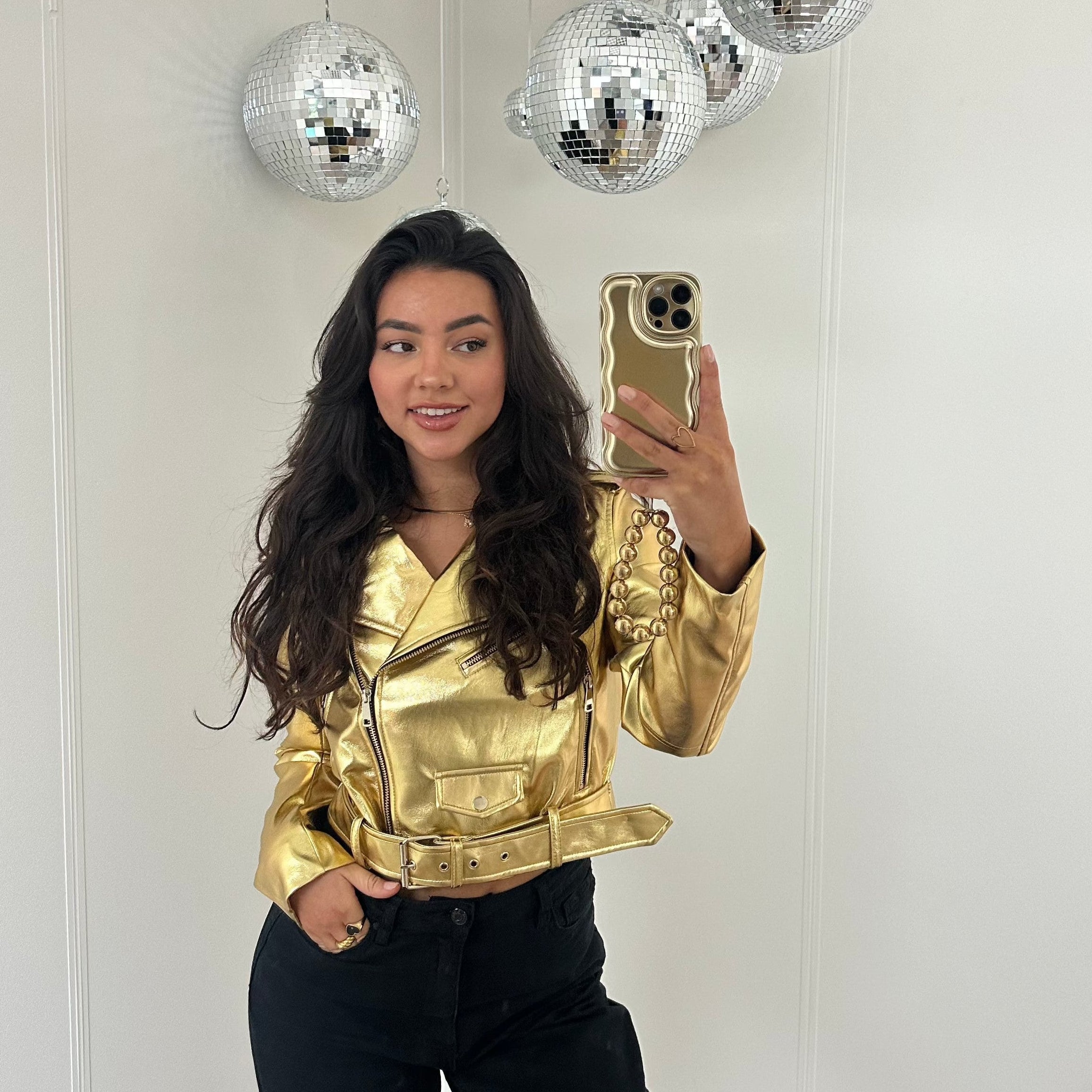 Young woman in a gold metallic biker jacket with a belted waist, holding a phone with a gold chain.